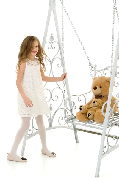 Blondýny dívka v elegantní bílé šaty houpající se její medvídek na houpačce. — Stock fotografie