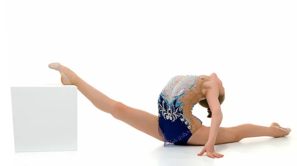 Beautiful girl gymnast on a white cube does an acrobatic twine element. — Stock Photo, Image