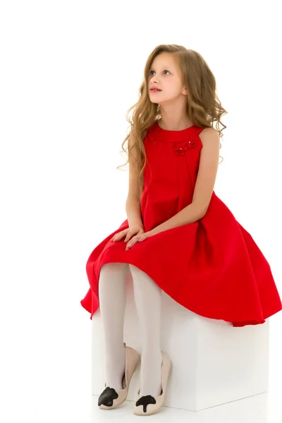 Menina loira encantadora em vestido vermelho sentado no cubo branco Olhando — Fotografia de Stock