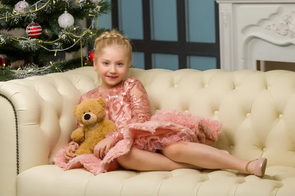 Een klein meisje met een teddybeer zit op een bank naast een kerstboom.. — Stockfoto