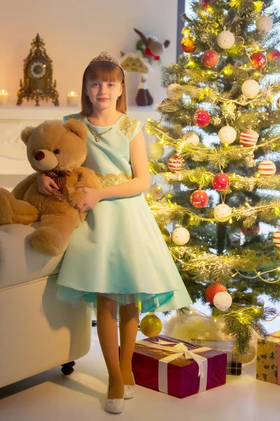Uma menina com um ursinho de pelúcia está sentada em um sofá perto de uma árvore de Natal. — Fotografia de Stock