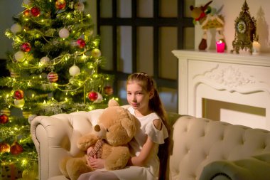 Oyuncak ayılı küçük bir kız Noel ağacının yanındaki kanepede oturuyor..