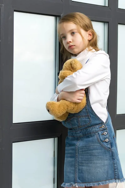 Uma menina fica perto da janela e abraça um ursinho de pelúcia. — Fotografia de Stock