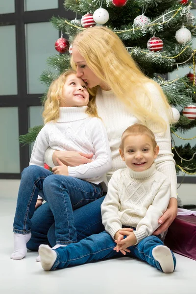 Uśmiechnięta mama przytula swoje dzieci przed dekorowaną choinką. — Zdjęcie stockowe