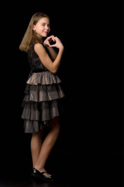 Küçük kız elleriyle kalp yapıyor ve siyah arka planda gülümsüyor..