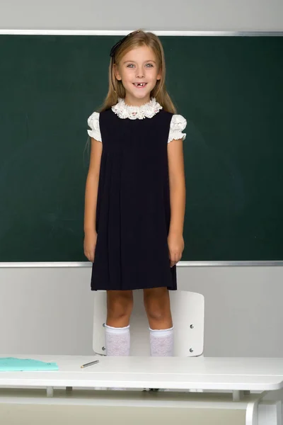 Estudante loira sorridente em uniforme escolar fica em uma cadeira na frente do quadro. Conceito de escola e educação — Fotografia de Stock