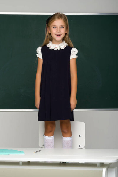 Smiling blonde female student in school uniform stands on a chair in front of the blackboard. School and education concept