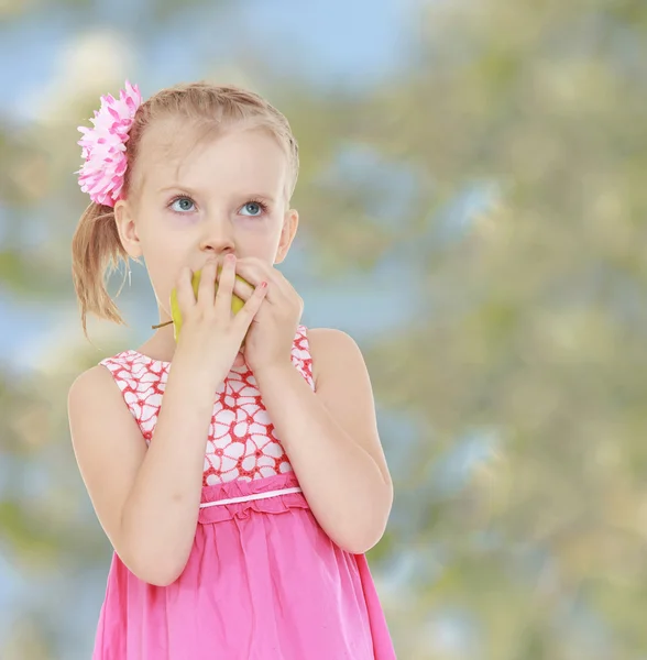 Liten flicka i en rosa klänning biter en apple, blekt gröna bak — Stockfoto