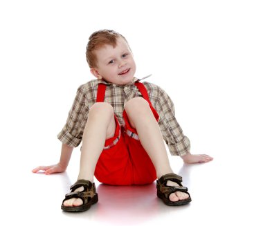 Joyful little boy in a plaid shirt, red shorts and flip-flops si