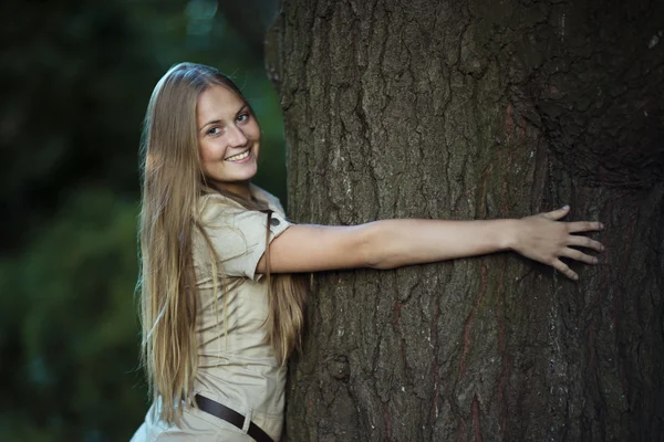 Joven chica atractiva abrazando un gran árbol en el parque y sonriendo —  Fotos de Stock