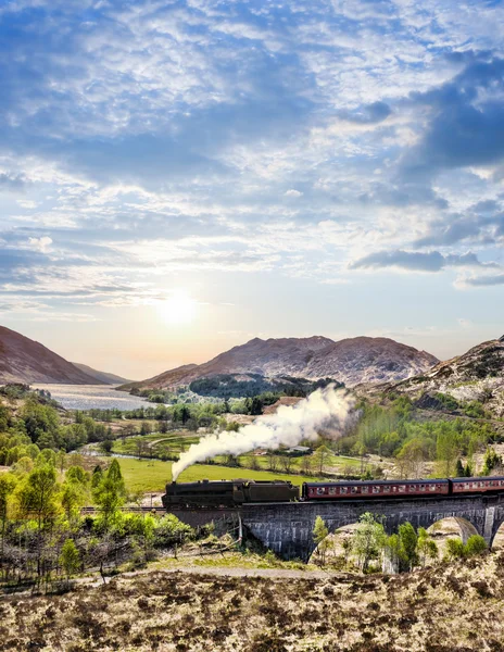 Glenfinnan Viaduto ferroviário na Escócia com o trem a vapor jacobita contra o pôr do sol sobre o lago — Fotografia de Stock