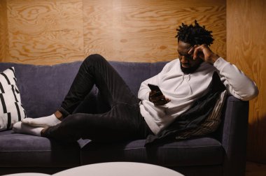 Gülümseyen Afrikalı Amerikalı adam evinde rahat bir koltukta oturup modern cep telefonuyla rahatlıyor. Melez erkek milenyumu koltukta dinleniyor. Oturma odasında akıllı telefon cihazına mesaj yazıyor.