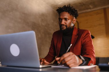 Odaklanmış Afrikalı genç işadamı kulaklık takıyor internetten internetten internetten internetten internetten internet yayınları izliyor eğitim dersleri konferansını dinliyor notlar alıyor çalışma masasında oturuyor, konsept öğreniyor.