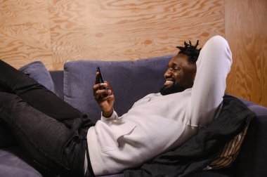 Gülümseyen Afrikalı Amerikalı adam evinde rahat bir koltukta oturup modern cep telefonuyla rahatlıyor. Melez erkek milenyumu koltukta dinleniyor. Oturma odasında akıllı telefon cihazına mesaj yazıyor.