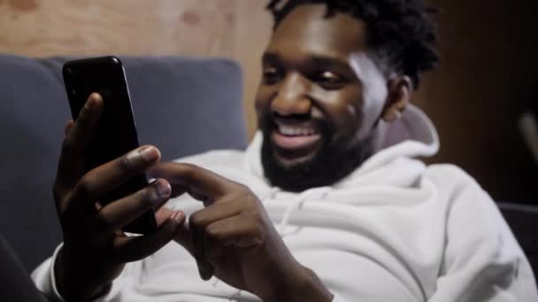 Sonriente hombre afroamericano sentarse relajarse en cómodo sofá en casa utilizando el teléfono celular moderno, feliz biracial milenario descanso masculino en el sofá en la sala de mensajes de texto de mensajería en el gadget smartphone — Vídeo de stock