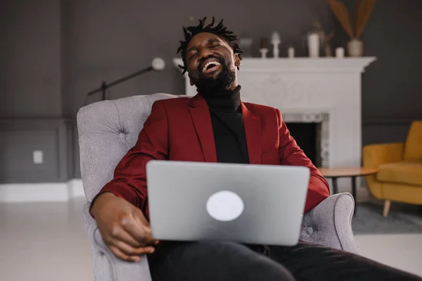 Um jovem afro-americano bonito e sorridente sentado, a olhar para o ecrã do computador. Homem biracial milenar focado trabalhando no projeto no escritório moderno ou estudando on-line em casa. — Fotografia de Stock