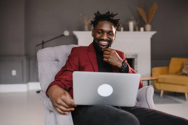 Um jovem afro-americano bonito e sorridente sentado, a olhar para o ecrã do computador. Homem biracial milenar focado trabalhando no projeto no escritório moderno ou estudando on-line em casa. — Fotografia de Stock