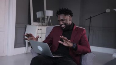 Gülümseyen, yakışıklı, Afro-Amerikan bir adam oturuyor ve bilgisayar ekranına bakıyor. Modern ofiste projeler üzerinde çalışan ya da evde internetten eğitim gören iki ırklı bir adam..
