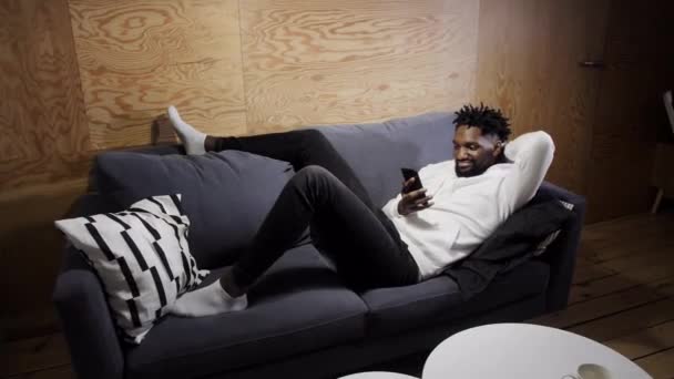 Черный мужчина отдыхает на диване и смотрит на телефон — стоковое видео