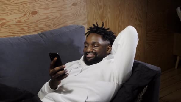 笑着的非洲裔美国男人坐在舒适的沙发上悠闲自在地使用现代手机，快乐而又千年的男性在客厅的沙发上休息，用智能手机工具发短信 — 图库视频影像