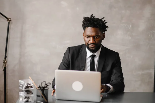 Snygg afrikansk amerikansk affärsman i klassisk kostym använder laptop och ler när du arbetar på kontoret — Stockfoto