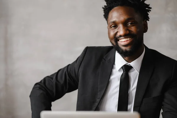 Snygg afrikansk amerikansk affärsman i klassisk kostym använder laptop och ler när du arbetar på kontoret — Stockfoto