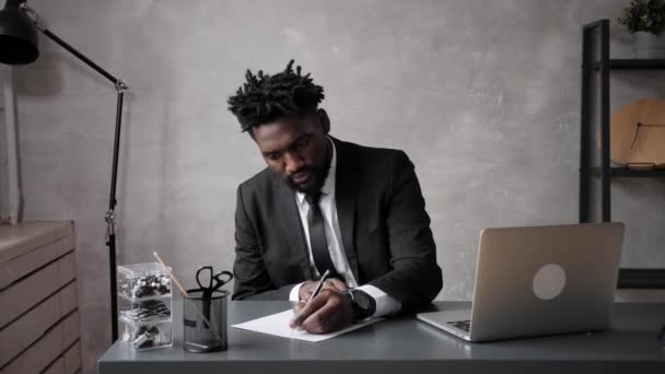 Een zwarte zakenman in een pak maakt aantekeningen op een vel papier. E-learning op afstand via een computer — Stockvideo