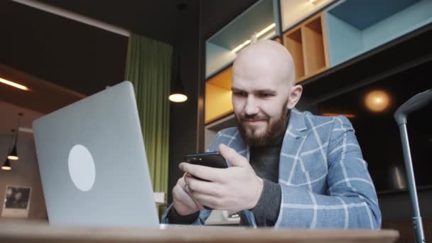 Un om de afaceri chel într-un costum care stă într-o cafenea lucrează la un laptop și navighează la telefon — Videoclip de stoc