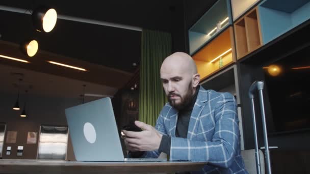 Hombre de negocios calvo en un traje sentado en un café trabaja en un ordenador portátil y navega por el teléfono — Vídeo de stock