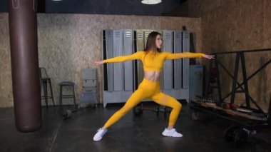 Sarı eşofmanlı güzel bir kız esneme hareketleri yapıyor. Geniş pencereli modern spor salonu