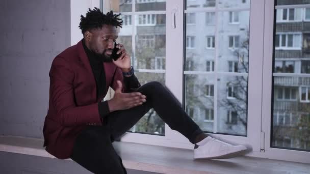 Souriant beau jeune homme afro-américain assis et parlant au téléphone. Homme biracial millénaire concentré travaillant sur un projet dans un bureau moderne ou étudiant en ligne à la maison. — Video