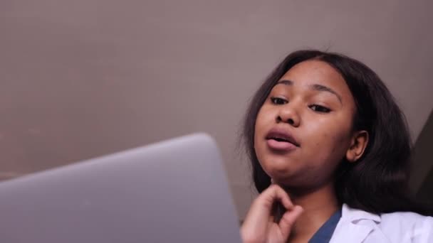 Femme amicale clinicienne noire en robe blanche en utilisant un ordinateur portable pour la consultation à distance des patients dans le bureau de l'hôpital. Télémédecine. Rendez-vous médecin en ligne. — Video