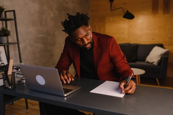 Homem negro afro-americano bonito trabalhando no computador portátil enquanto sentado atrás da mesa na sala de estar aconchegante. Freelancer trabalhando em casa — Fotografia de Stock