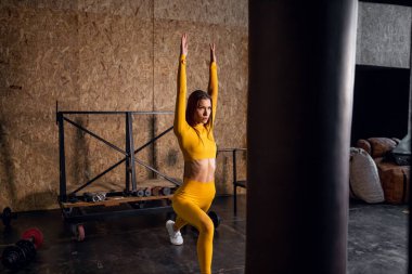 Sarı eşofmanlı güzel bir kız esneme hareketleri yapıyor. Geniş pencereli modern spor salonu