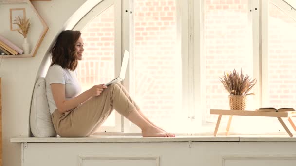 A young woman sits on the windowsill in a cozy bright apartment and communicates via video. attractive woman uses laptop for her work — Stock Video