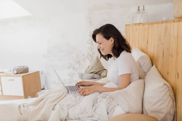 Gelukkig mooi vrouw werken op een laptop terwijl zitten op een bed in een helder modern huis. — Stockfoto