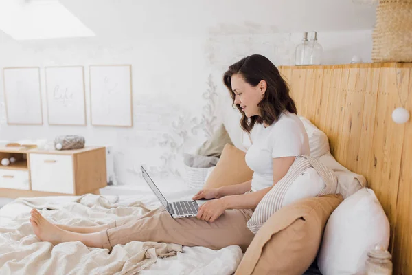 Ευτυχισμένη όμορφη γυναίκα που εργάζεται σε ένα φορητό υπολογιστή, ενώ κάθεται σε ένα κρεβάτι σε ένα φωτεινό σύγχρονο σπίτι. — Φωτογραφία Αρχείου