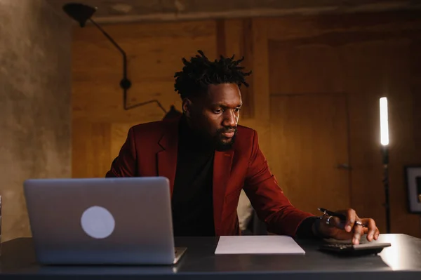 Homem negro afro-americano bonito trabalhando no computador portátil enquanto sentado atrás da mesa na sala de estar aconchegante. Freelancer trabalhando em casa — Fotografia de Stock