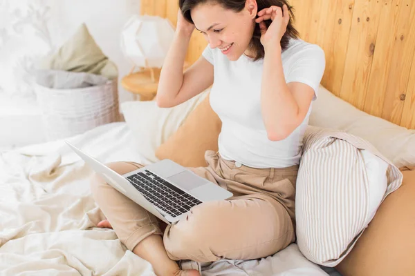 Ung attraktiv kvinna ligger på en säng i arbetar på en bärbar dator. Fjärrarbete hemifrån. E-lärande — Stockfoto