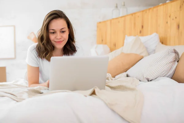 Una mujer joven se acuesta en la cama y utiliza un ordenador portátil para videoconferencias o charlar con amigos — Foto de Stock