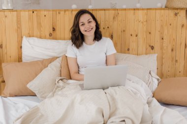 Genç ve çekici bir kadın dizüstü bilgisayarla yatakta yatıyor. Evden uzak bir iş. E-öğrenme