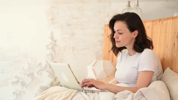Jovem mulher atraente deitado em uma cama em trabalhar em um laptop. Trabalho remoto de casa. E-learning — Vídeo de Stock