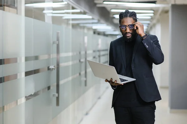 Retrato de um afro-americano num fato preto no fundo do escritório. homem de negócios preto com um laptop — Fotografia de Stock