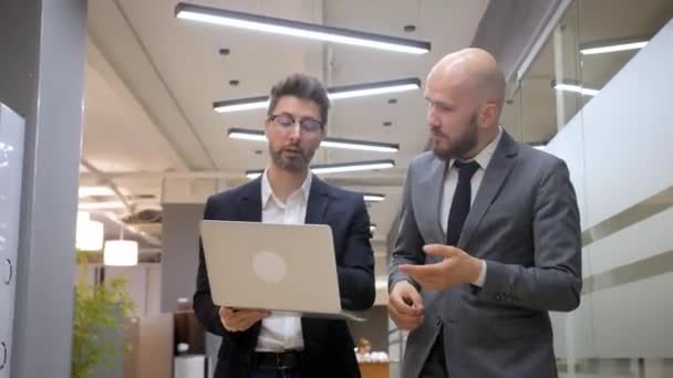 Dois empresários caminham pelo corredor do escritório até uma reunião. Eles estão discutindo um novo projeto em um laptop. — Vídeo de Stock