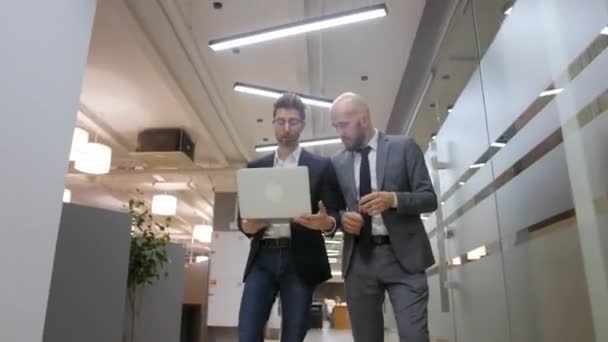 Dois empresários caminham pelo corredor do escritório até uma reunião. Eles estão discutindo um novo projeto em um laptop. — Vídeo de Stock