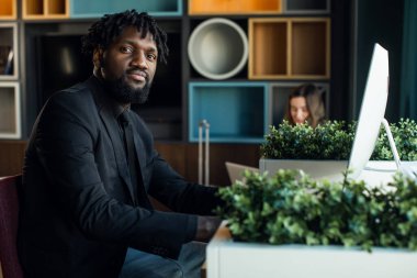 Koyu renk takım elbiseli esmer iş adamı modern bir ofiste bilgisayar başında çalışıyor.