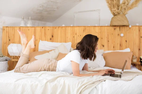 Νεαρή ελκυστική κοπέλα, χρησιμοποιεί ένα φορητό υπολογιστή, ενώ στο κρεβάτι. Μακρινή εργασία από το σπίτι — Φωτογραφία Αρχείου