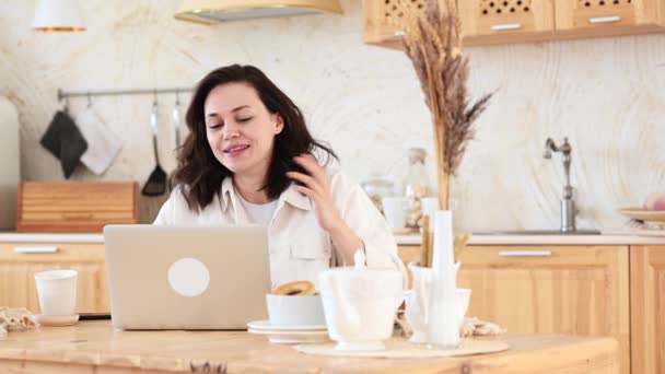 Een jonge aantrekkelijke vrouw zit aan een tafel in haar keuken en praat door middel van een video conferentie — Stockvideo