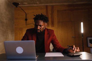 Takım elbiseli odaklanmış siyah bir adam dizüstü bilgisayar üzerinde çalışıyor. Evden uzak çalışma