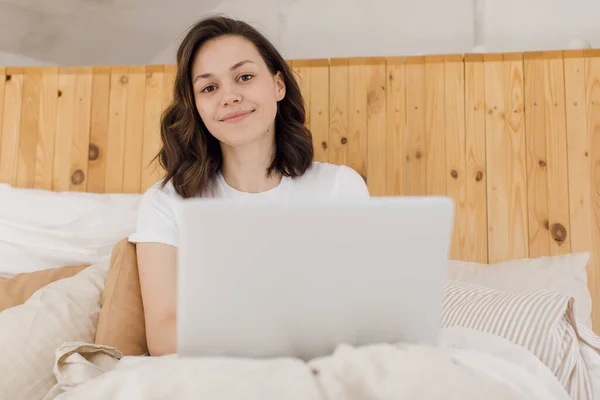 Ung attraktiv kvinna ligger på en säng i arbetar på en bärbar dator. Fjärrarbete hemifrån. E-lärande — Stockfoto